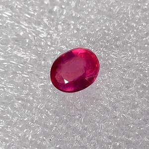 Puede incluir: Una gema ovalada de color rojo vibrante, probablemente un rubí, se muestra sobre un fondo blanco texturizado.