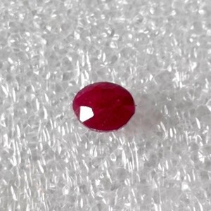 Puede incluir: Una piedra preciosa ovalada de color rojo oscuro, probablemente un rubí, sobre un fondo blanco texturizado.