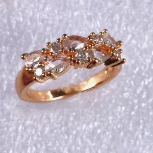 Puede incluir: Un anillo dorado con un grupo de piedras preciosas ovaladas y redondas. La banda del anillo es de color oro cálido, y las piedras son una mezcla de tonos claros y melocotón claro, creando un efecto brillante.