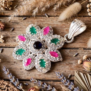May include: A silver pendant shaped like a flower, featuring a central dark blue stone surrounded by alternating marquise-cut green and pink stones, all accented with small clear pavé crystals and a textured silver bail at the top.
