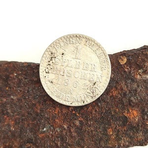 May include: A silver coin from 1861 with German text, resting on a piece of rusted metal. The coin features the number 1 and the words "SILBER GROSCHEN". The metal has a rough, textured surface and a dark brown color.