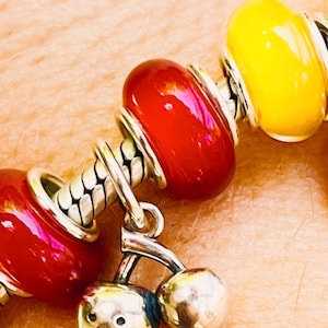 Può includere: Un braccialetto con ciondoli argentato con perline rosse e gialle. Il braccialetto presenta un ciondolo a forma di ananas argentato e un ciondolo a forma di ciliegia argentata. Le perline sono di un vivace colore rosso e giallo e il braccialetto è indossato al polso.