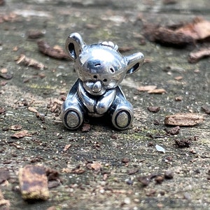 May include: A small, silver-coloured teddy bear charm in a sitting pose. The bear has a rounded head with small ears and a button nose. The charm is detailed with a textured surface and is suitable for jewellery making.