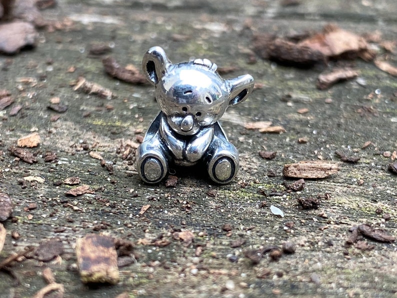 May include: A small, silver-coloured teddy bear charm in a sitting pose. The bear has a rounded head with small ears and a button nose. The charm is detailed with a textured surface and is suitable for jewellery making.