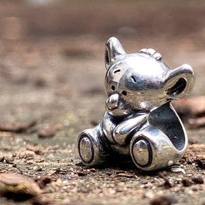 May include: A silver charm in the shape of a koala bear sitting down. The charm is detailed with a small nose and ears.
