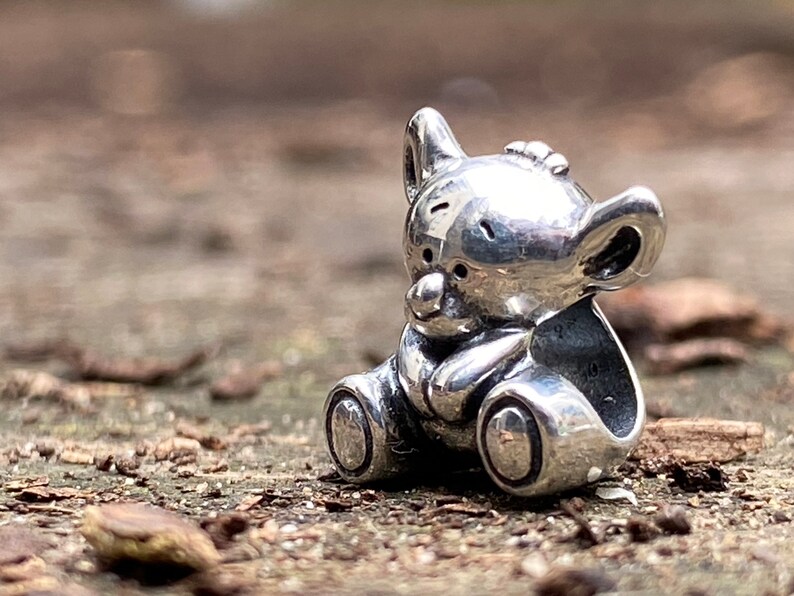 May include: A silver charm in the shape of a koala bear sitting down. The charm is detailed with a small nose and ears.
