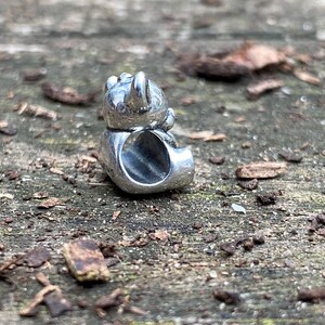 May include: Silver charm in the shape of a cartoon bear sitting down. The bear is wearing a hat and has a small hole in the back for attaching to a bracelet.