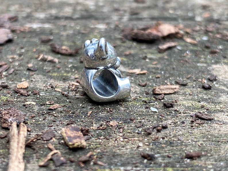 May include: Silver charm in the shape of a cartoon bear sitting down. The bear is wearing a hat and has a small hole in the back for attaching to a bracelet.