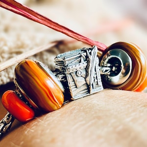 May include: A silver charm bracelet featuring a suitcase-shaped charm with a feather on top. The bracelet also includes three glass beads, one red, one orange, and one brown with orange stripes.