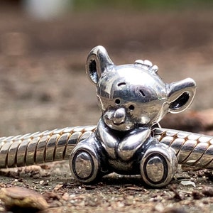 May include: A silver charm in the shape of a teddy bear. The bear is sitting with its paws in front of it. The charm is designed to be used on a bracelet or necklace.