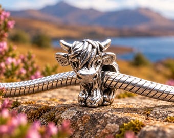 May include: A polished silver charm shaped like a Highland cow with detailed fur texture, horns, and ears, threaded onto a flexible silver snake chain bracelet. The charm features a front-facing cow head with a shaggy fringe and small hooves.