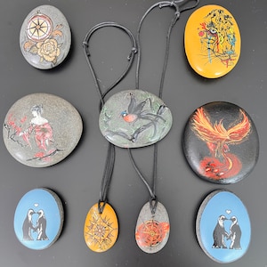 Pode incluir: Uma coleção de dez pedras pintadas à mão com vários desenhos. As pedras são pintadas com uma variedade de cores e padrões, incluindo uma bússola, um pássaro, uma fênix, um leopardo e pinguins. Algumas das pedras são enfiadas com um cordão preto.