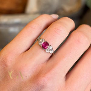 Könnte beinhalten: Ein goldener Ring mit einem großen ovalen Rubin in der Mitte, flankiert von zwei Gruppen kleiner, runder, weißer Edelsteine. Der zarte Ringreif unterstreicht das klassische Design. Die Edelsteine funkeln im Kontrast zum Gold. Ein zeitloses Schmuckstück.