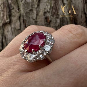 May include: A silver ring featuring a large, oval-cut ruby surrounded by a halo of smaller, round diamonds. The ruby is a deep red color, and the diamonds sparkle brightly. The ring is set against a natural background.