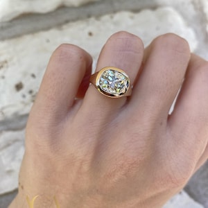 May include: A gold ring with a large, cushion-cut diamond. The diamond is set in a bezel setting, and the ring has a smooth, polished band. The ring is shown on a hand, with a brick wall in the background.
