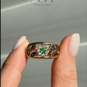 May include: A gold ring featuring a floral design with a central emerald surrounded by diamonds. The ring is further adorned with ruby, sapphire, and emerald accents. The ring is held up by a person's fingers.