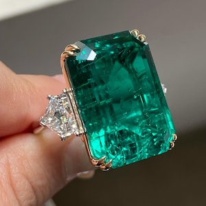 May include: A striking emerald ring. The ring features a large, rectangular emerald, set in a gold bezel, and two smaller, triangular diamonds. The emerald is a deep green colour. The setting is gold and silver. The ring is a statement piece.