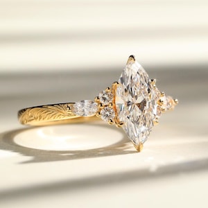 Op de afbeelding: Een gouden verlovingsring met een marquise-geslepen diamant als middelpunt. De ring heeft kleinere accentdiamanten en een gedetailleerd bloemmotief op de band. De ring ligt op een wit oppervlak.