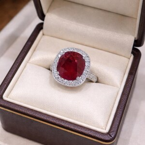 Könnte beinhalten: Ein Ring mit einem kissenförmigen Rubin, umgeben von einem Halo aus kleinen Diamanten, präsentiert in einer braunen Schmuckschatulle. Der Ring hat ein silberfarbenes Band, der Rubin ist tiefrot.