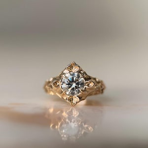 May include: A gold ring featuring a central round brilliant-cut clear gemstone set within a diamond-shaped frame. The metal band displays intricate, raised floral-inspired carvings that extend along the sides of the setting, creating a textured surface.