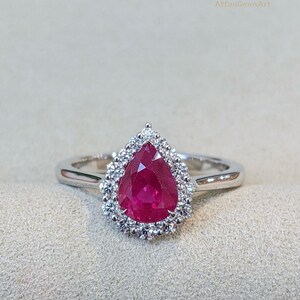 Puede incluir: Un anillo de plata con una piedra preciosa roja oscura en forma de pera, rodeada por un halo de piedras claras más pequeñas. La banda del anillo es fina y pulida, y el engaste resalta la piedra central. El texto "AGA AffanGemsArt" está en la esquina superior derecha.