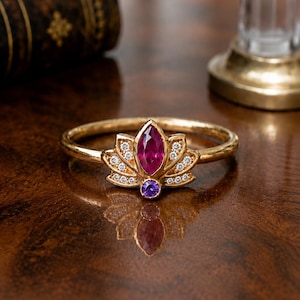 May include: A gold ring featuring a lotus flower design. The ring has a large, oval-shaped ruby gemstone at the center, with a smaller, round purple gemstone below. Small diamonds are set around the ruby. The ring is on a wooden surface.