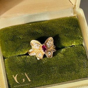 May include: A gold butterfly ring with a central oval ruby and small clear gemstones. The ring is displayed in a vintage green velvet lined jewellery box. The box is cream coloured with the text "JEWELLERS TORONTO".
