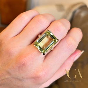May include: A large, rectangular-cut, light green gemstone ring. The ring features a silver band and a prominent, emerald-cut stone. The gemstone has a translucent quality, with visible internal structures. The ring is set on a hand, showcasing its size and color.