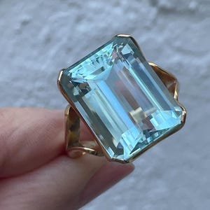 May include: A gold ring with a large, rectangular, light blue gemstone. The gemstone is faceted, reflecting light. The ring's band has an open design. The ring is held against a light grey background. The gemstone is approximately 2.5 cm long.