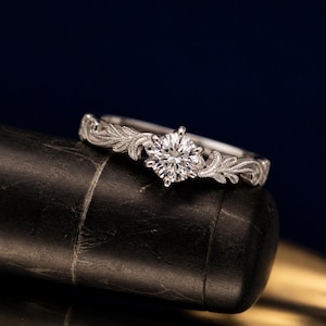 May include: A silver ring with a large, round, clear gemstone at the centre. The band features a delicate, leaf-like pattern. The ring is displayed on a dark, textured surface against a deep blue background.
