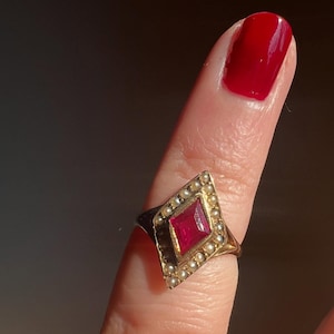 Könnte beinhalten: Ein goldener Ring mit rautenförmiger Fassung. Die Fassung hat einen großen, rechteckigen, roten Edelstein, der von kleinen, runden Perlen umgeben ist. Der Ring befindet sich an einem Finger mit rotem Nagellack.