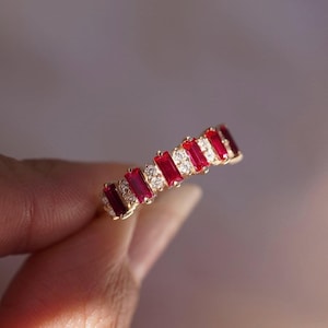 Può includere: Un anello d'oro con pietre rosse rettangolari alternate e piccole pietre chiare rotonde. L'anello è tenuto su uno sfondo morbido e sfocato, evidenziando il design e la brillantezza del gioiello.