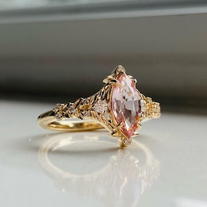 May include: A gold ring with a light pink marquise-cut gemstone. The ring has a floral design with small, clear accent stones. The band and setting are gold, reflecting light. The ring is set against a white surface.