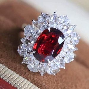 Könnte beinhalten: Silberring mit einem großen, oval geschliffenen, tiefroten Edelstein, umgeben von einem Kranz aus kleineren, klaren, birnenförmigen und runden Edelsteinen. Der Ring ist vor einem neutralen Hintergrund platziert.