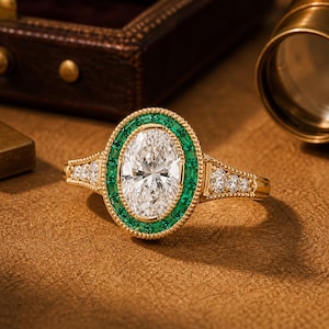 May include: A gold ring featuring an oval diamond centre stone, encircled by emeralds. The band is adorned with small diamonds and detailed patterns. The ring is displayed on a warm, brown surface.