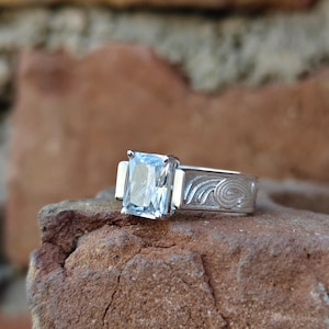 Könnte beinhalten: Ein silberner Ring mit einem großen, rechteckigen, hellblauen Edelstein. Das Ringband weist ein graviertes Wirbeldesign auf. Der Ring ist auf einem rauen, braunen Stein platziert, mit einem verschwommenen Ziegelhintergrund.