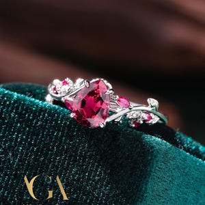 Könnte beinhalten: Ein silberner Ring mit einem großen, birnenförmigen, tiefroten Edelstein. Der Ring hat kleinere rote Edelsteine und blattartige Details. Der Ring ist vor einem dunkelgrünen Hintergrund platziert.
