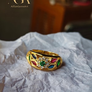 May include: A gold ring with a central cluster of diamonds and a green gemstone, surrounded by colorful gemstones in red, blue, and green. The ring is set on a white, textured surface.