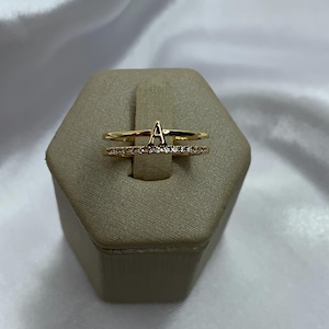Puede incluir: Un conjunto de anillos dorados. El anillo superior presenta la letra "A" en escritura cursiva. El anillo inferior está adornado con pequeñas piedras preciosas transparentes. Los anillos se exhiben sobre un expositor de joyas hexagonal beige sobre un fondo blanco.