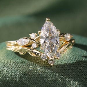 May include: A gold engagement ring set with a large, marquise-cut diamond as the centerpiece. The band features smaller diamonds and a delicate, leaf-like design. The ring is set against a green background.