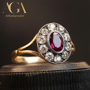 May include: A gold ring with an oval ruby centre stone, encircled by a halo of diamonds. The ring's design includes a split shank. The brand name "AffanGemsArt" is displayed in the upper left corner against a dark backdrop.