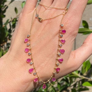 May include: A gold necklace with a series of pink teardrop-shaped gemstones and small, clear, round gemstones. The necklace is shown against a natural green background.