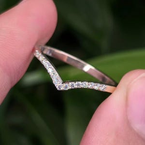 May include: A V-shaped ring crafted from a polished silver-toned metal, featuring a row of small, clear, round-cut stones embedded along the top surface of the band, creating a continuous sparkling effect across the chevron-style design.