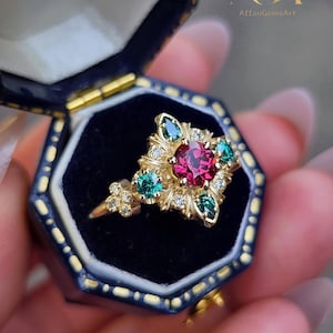 Puede incluir: Un anillo de oro con una piedra preciosa roja central, rodeada de piedras preciosas verdes y azules más pequeñas y piedras de acento transparentes. El anillo se muestra en un joyero forrado de terciopelo azul. El diseño del anillo es ornamentado y detallado.