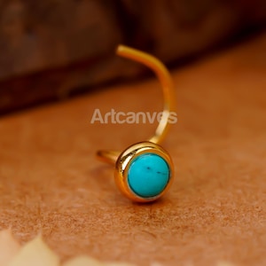 May include: A gold nose ring featuring a small, round turquoise stone in a bezel setting. The ring has a curved post and a circular setting. The background is a warm, neutral tone, with the text "Artcanvs" visible.