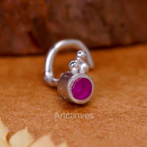 Puede incluir: Un aro de nariz de plata con un engaste circular y una piedra de color rosa vibrante. La pieza de joyería incluye un pequeño grupo de tres esferas de plata situadas sobre el engaste, unidas a un poste de alambre curvo para su inserción.