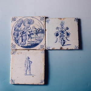 Può includere: Quattro antiche piastrelle in ceramica con disegni blu e bianchi. Una piastrella presenta una scena con figure, un'altra una composizione floreale e una terza raffigura un soldato. Le piastrelle hanno motivi decorativi agli angoli.
