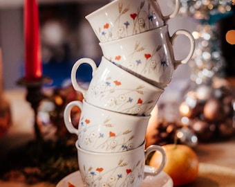 Peut inclure: Une pile de cinq tasses à thé blanches avec des motifs floraux et de blé, empilées les unes sur les autres. Les tasses sont sur un ensemble de soucoupes blanches, avec une petite barre chocolatée brune et blanche sur le dessus des soucoupes. Les tasses et les soucoupes sont sur un napperon tissé.