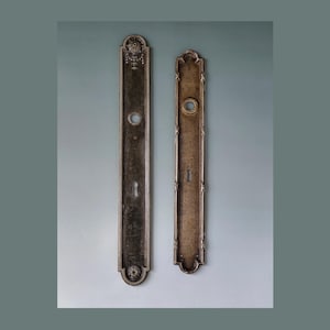 Può includere: Due antiche placche per porte in bronzo con dettagli ornati. Le placche sono rettangolari con bordi decorativi, fori per chiavi e accenti circolari. Una placca ha un motivo floreale in alto e in basso, l'altra ha un pomello circolare.