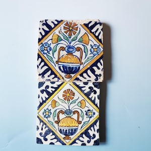 Può includere: Due piastrelle quadrate in ceramica con un motivo floreale. Ogni piastrella presenta un vaso centrale con fiori gialli, arancioni e blu, all'interno di una forma a diamante. Il diamante è bordato da un motivo giallo e blu. Le piastrelle sono su uno sfondo azzurro.